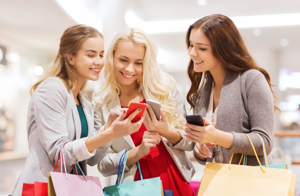 Three happy young women with shopping bags in mall looking at their smartphones